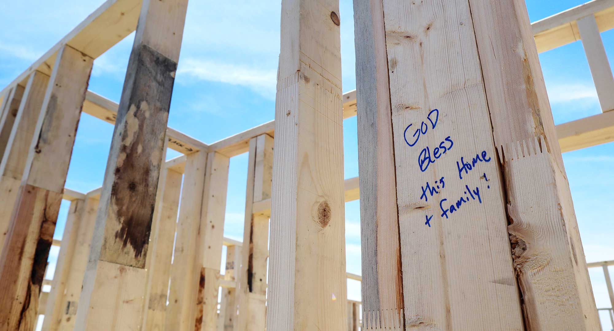 Framed walls of a home with "God bless this home + family!" written on a 2x4